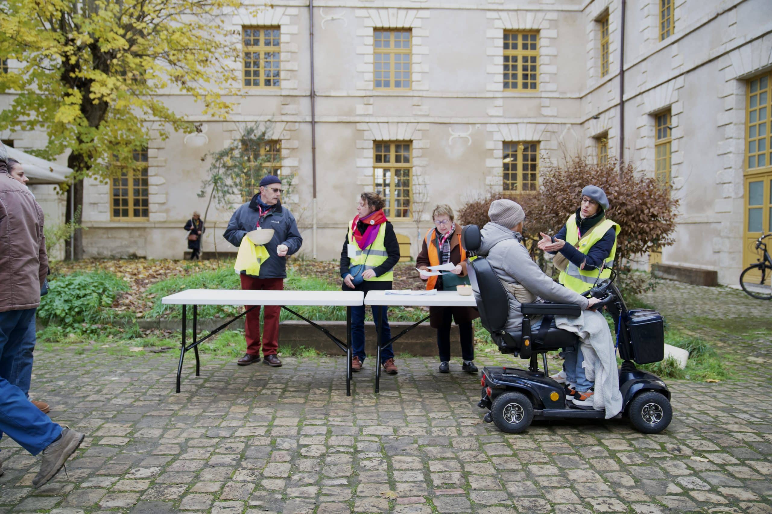 Des bénévoles en gilets jaunes discutent avec une personne en fauteuil roulant électrique devant une table d'accueil installée dans la cour d'un bâtiment historique lors du lancement du LICIA à Bourges.